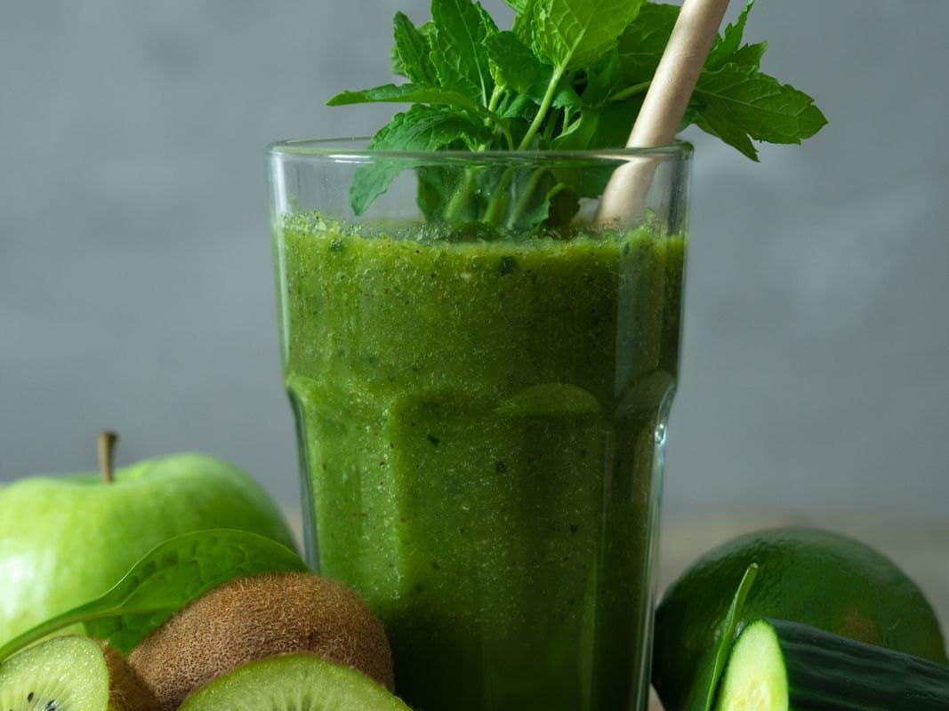 Düz bardakta koyu yeşil smoothie, üzerinde nane demeti, yanında yeşil elma ve kivi