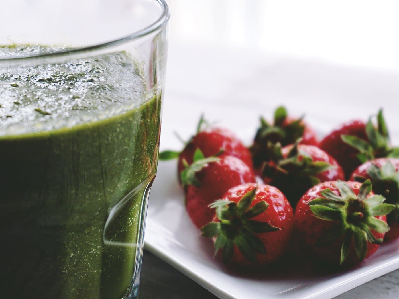 Bardakta koyu yeşil ıspanaklı smoothie, yanında beyaz tabakta bütün çilekler